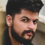 Close-up portrait of a South Asian man with a mustache and beard, dressed in black, outdoors.